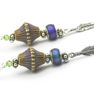 Pu&ograve; includere: Un paio di orecchini pendenti con un ciondolo a forma di piuma d'argento, una perla blu iridescente e una perla marrone e viola con un motivo.