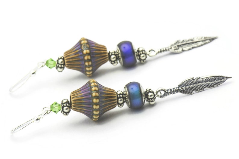 Pu&ograve; includere: Un paio di orecchini pendenti con un ciondolo a forma di piuma d'argento, una perla blu iridescente e una perla marrone e viola con un motivo.