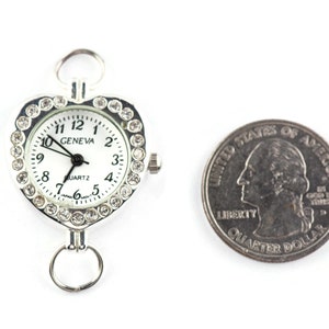 Pu&ograve; includere: Un orologio a forma di cuore argentato con un quadrante bianco e accenti di strass trasparenti. Il quadrante dell'orologio recita "GENEVA QUARTZ" e ha lancette nere. L'orologio &egrave; accanto a un quarto di dollaro americano per il confronto delle dimensioni.