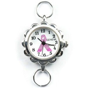 May include: A silver watch with a white face and a pink ribbon design. The watch has a Geneva Quartz movement and is made in Japan. The watch is attached to a silver chain with a ring at each end.