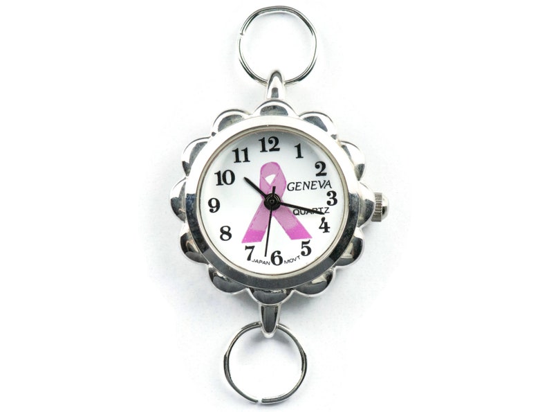 May include: A silver watch with a white face and a pink ribbon design. The watch has a Geneva Quartz movement and is made in Japan. The watch is attached to a silver chain with a ring at each end.