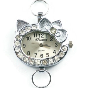 May include: A silver charm watch with a white face and rhinestones around the edge. The watch face has black numbers and the word "Dagicci" printed on it. The watch has a silver bow on top and a silver loop on the bottom.