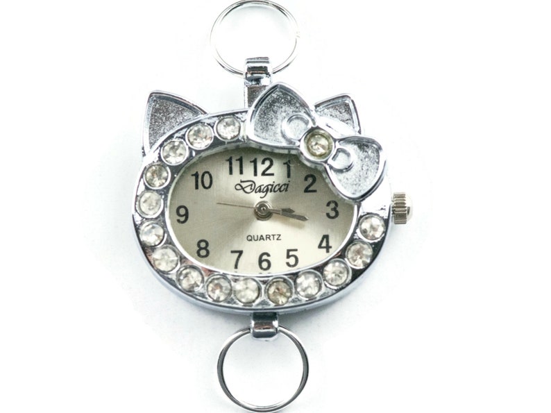 May include: A silver charm watch with a white face and rhinestones around the edge. The watch face has black numbers and the word "Dagicci" printed on it. The watch has a silver bow on top and a silver loop on the bottom.