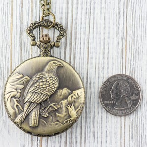 May include: A gold-toned pocket watch with a chain. The watch face features an engraved design of a bird perched on a branch. The watch is about the size of a US quarter.