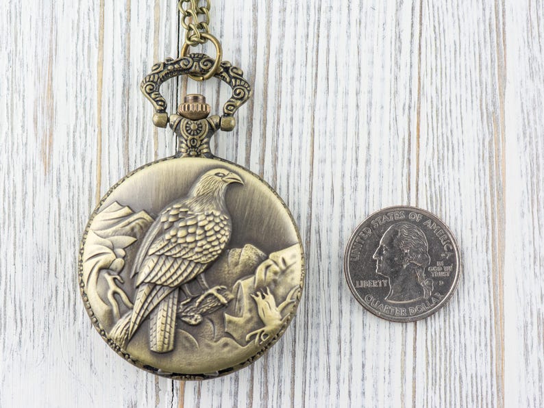 May include: A gold-toned pocket watch with a chain. The watch face features an engraved design of a bird perched on a branch. The watch is about the size of a US quarter.
