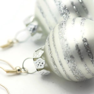 K&ouml;nnte beinhalten: Ein Paar Ohrringe aus silbernen und wei&szlig;en Weihnachtskugeln. Die Kugeln sind rund und haben eine silberne Glitzer-Oberfl&auml;che. Die Ohrringe sind an silbernen Haken befestigt.