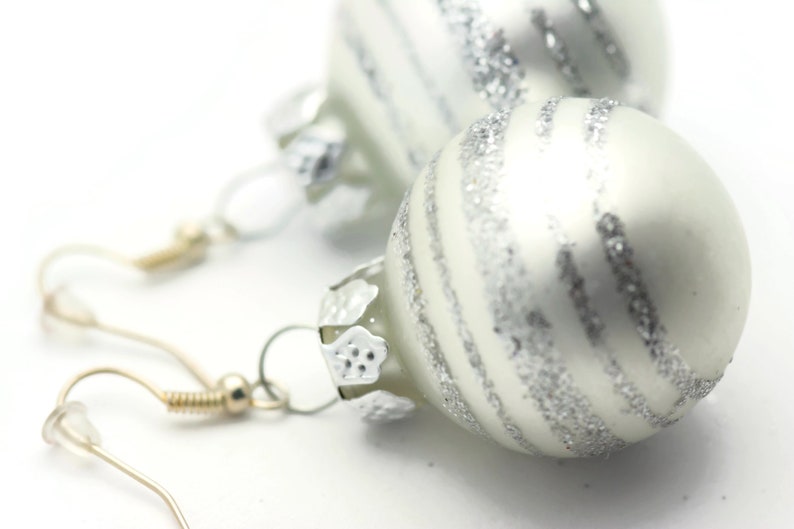 K&ouml;nnte beinhalten: Ein Paar Ohrringe aus silbernen und wei&szlig;en Weihnachtskugeln. Die Kugeln sind rund und haben eine silberne Glitzer-Oberfl&auml;che. Die Ohrringe sind an silbernen Haken befestigt.