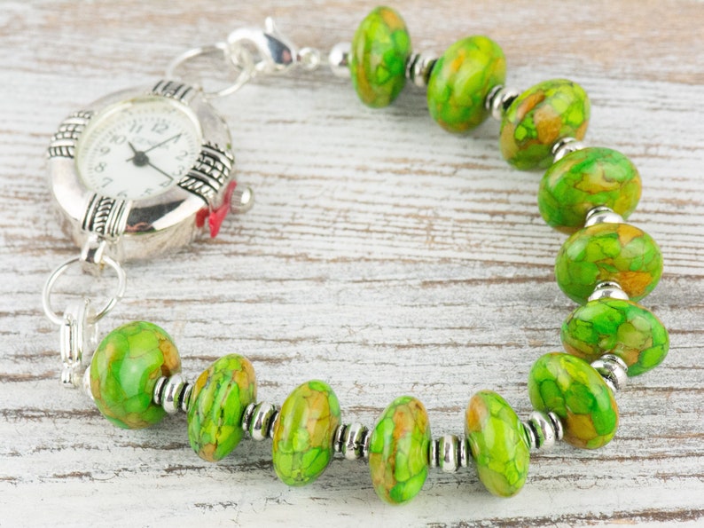 Pode incluir: Um rel&oacute;gio prateado com uma pulseira de contas com padr&atilde;o verde e marrom. O mostrador do rel&oacute;gio tem ponteiros prateados e n&uacute;meros pretos.