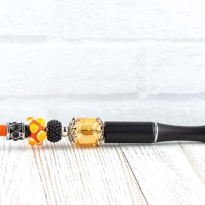May include: A black pen with decorative beads. The beads include orange, yellow, and black designs. The pen has a silver tip and a black barrel. The pen is on a white wooden surface.