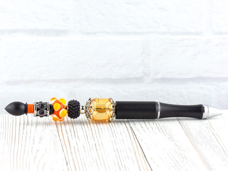 May include: A black pen with decorative beads. The beads include orange, yellow, and black designs. The pen has a silver tip and a black barrel. The pen is on a white wooden surface.