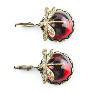 Pode incluir: Um par de brincos com uma pedra de caboch&atilde;o vermelha e preta e um design de lib&eacute;lula dourada.