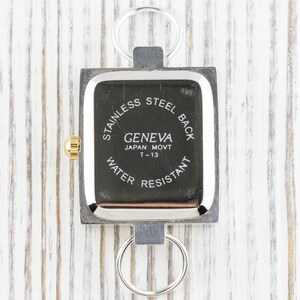 Peut inclure: Une montre Geneva carr&eacute;e avec un cadran noir et un cadre en m&eacute;tal argent&eacute;. La montre a une couronne dor&eacute;e et deux anneaux argent&eacute;s attach&eacute;s en haut et en bas. Le cadran indique "STAINLESS STEEL BACK", "GENEVA JAPAN MOVT T-13 WATER RESISTANT".