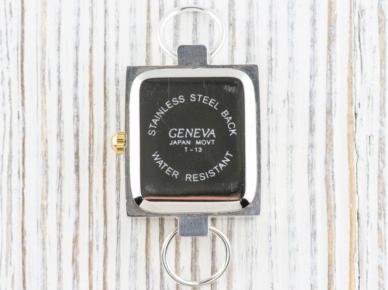 Peut inclure: Une montre Geneva carr&eacute;e avec un cadran noir et un cadre en m&eacute;tal argent&eacute;. La montre a une couronne dor&eacute;e et deux anneaux argent&eacute;s attach&eacute;s en haut et en bas. Le cadran indique "STAINLESS STEEL BACK", "GENEVA JAPAN MOVT T-13 WATER RESISTANT".