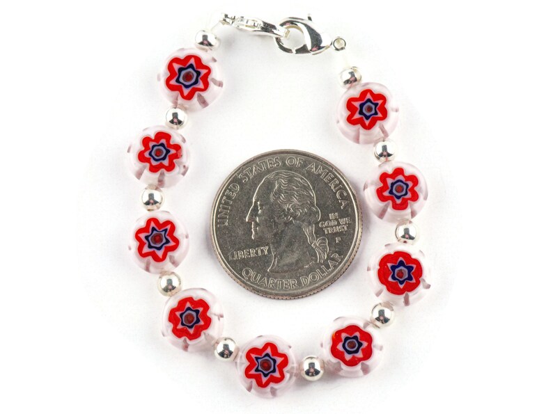 Puede incluir: Un brazalete de plata con cuentas de vidrio blancas y rojas con un dise&ntilde;o de estrella. El brazalete es aproximadamente del tama&ntilde;o de una moneda de 25 centavos de d&oacute;lar estadounidense.