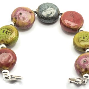 May include: A bracelet made of ceramic beads in shades of green, red, and grey. The beads are speckled with different colours and have a glossy finish. The bracelet is secured with a silver clasp.
