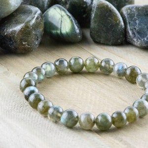 Puede incluir: Un brazalete de cuentas de labradorita con piedras redondas y pulidas. Las cuentas son de color verde claro con destellos azules. El brazalete se exhibe sobre una superficie de madera clara, con piedras sueltas de labradorita en el fondo.