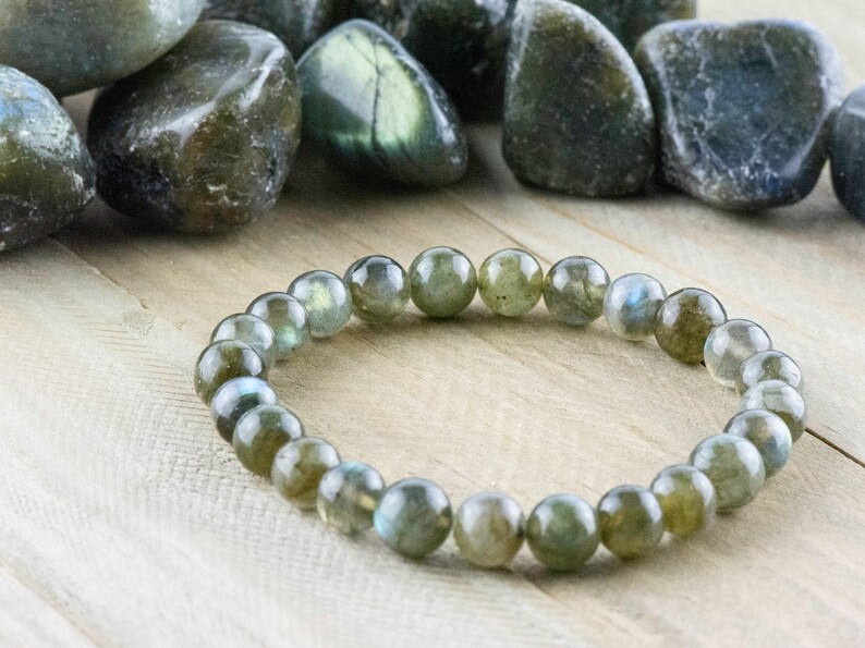 Puede incluir: Un brazalete de cuentas de labradorita con piedras redondas y pulidas. Las cuentas son de color verde claro con destellos azules. El brazalete se exhibe sobre una superficie de madera clara, con piedras sueltas de labradorita en el fondo.