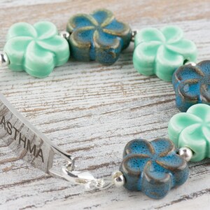 May include: A silver medical alert bracelet with a blue and green flower charm design. The bracelet has a metal tag that reads "ASTHMA" with a medical symbol.