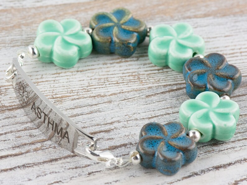 May include: A silver medical alert bracelet with a blue and green flower charm design. The bracelet has a metal tag that reads "ASTHMA" with a medical symbol.