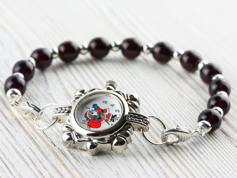 May include: A silver watch with a Santa Claus design on the face, attached to a bracelet made of dark red beads.