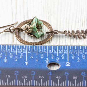 May include: A handcrafted earring with a turquoise stone, a bronze ring, and a fern charm. The earring is about 5 cm long, as measured by the ruler in the image. The earring has a hook closure and is made of metal.