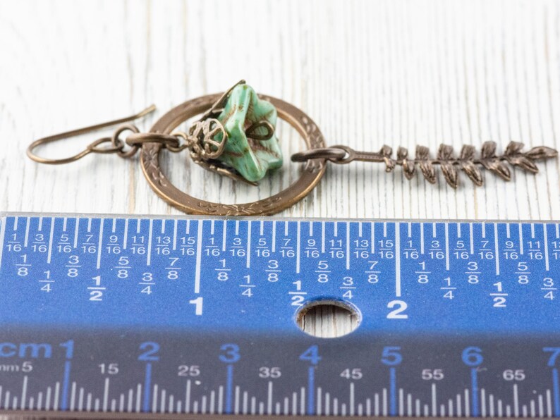 May include: A handcrafted earring with a turquoise stone, a bronze ring, and a fern charm. The earring is about 5 cm long, as measured by the ruler in the image. The earring has a hook closure and is made of metal.