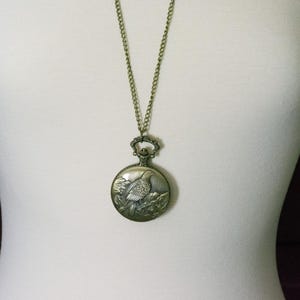 May include: A gold-toned necklace with a pocket watch pendant featuring an engraved image of a bird on a branch.