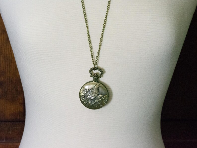 May include: A gold-toned necklace with a pocket watch pendant featuring an engraved image of a bird on a branch.