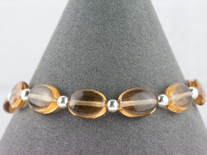 May include: A brown gemstone bracelet with silver beads. The bracelet is made of oval-shaped stones and is displayed on a gray background.