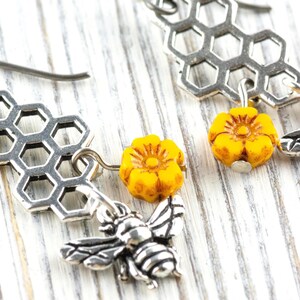 May include: Silver-coloured bee earrings with a honeycomb design and yellow flower accents. The earrings feature detailed bee charms and small yellow flower beads. The earrings are designed to dangle and are set against a white wooden background.