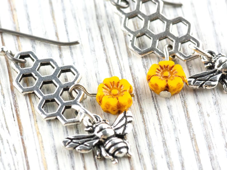 May include: Silver-coloured bee earrings with a honeycomb design and yellow flower accents. The earrings feature detailed bee charms and small yellow flower beads. The earrings are designed to dangle and are set against a white wooden background.