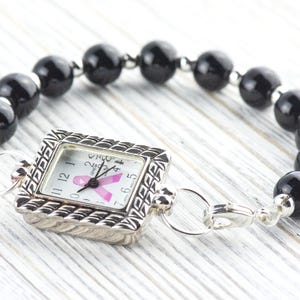 May include: A silver-toned watch with a square face and a black beaded bracelet. The watch face features a pink ribbon, and the bracelet is made of large, round black beads. The watch is connected to the bracelet with silver clasps.