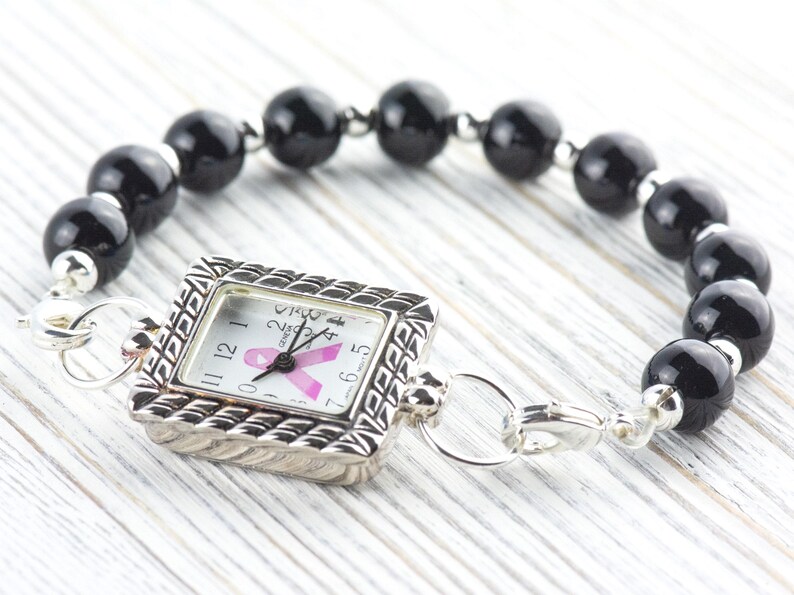 May include: A silver-toned watch with a square face and a black beaded bracelet. The watch face features a pink ribbon, and the bracelet is made of large, round black beads. The watch is connected to the bracelet with silver clasps.