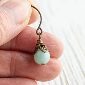 May include: Close-up of a single earring held by a person. The earring features a pale blue, teardrop-shaped bead suspended from a decorative bronze-colored cap and hook. The hook is a simple, curved wire.