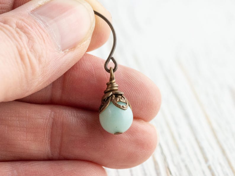 May include: Close-up of a single earring held by a person. The earring features a pale blue, teardrop-shaped bead suspended from a decorative bronze-colored cap and hook. The hook is a simple, curved wire.