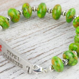 Pode incluir: Um bracelete de alerta m&eacute;dica verde e prateado com as palavras "Type I Diabetes" gravadas em uma placa de metal. O bracelete tem um s&iacute;mbolo m&eacute;dico e &eacute; feito com contas de pedra verde.