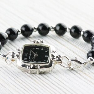 Pu&ograve; includere: Un orologio argentato con quadrante nero quadrato e bracciale di perline nere. Il quadrante dell'orologio ha numeri e lancette bianche. Il bracciale &egrave; composto da grandi perline nere lucide e perline argentate pi&ugrave; piccole. L'orologio &egrave; collegato al bracciale con chiusure argentate.