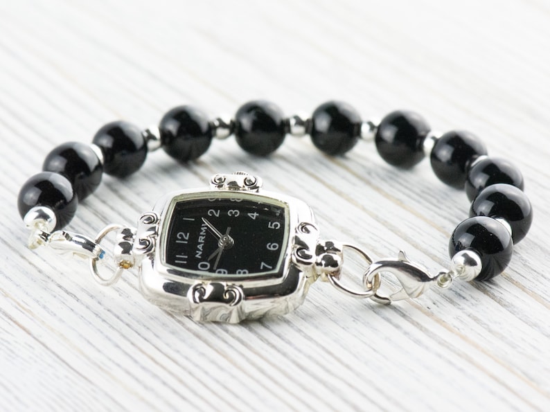 Pu&ograve; includere: Un orologio argentato con quadrante nero quadrato e bracciale di perline nere. Il quadrante dell'orologio ha numeri e lancette bianche. Il bracciale &egrave; composto da grandi perline nere lucide e perline argentate pi&ugrave; piccole. L'orologio &egrave; collegato al bracciale con chiusure argentate.