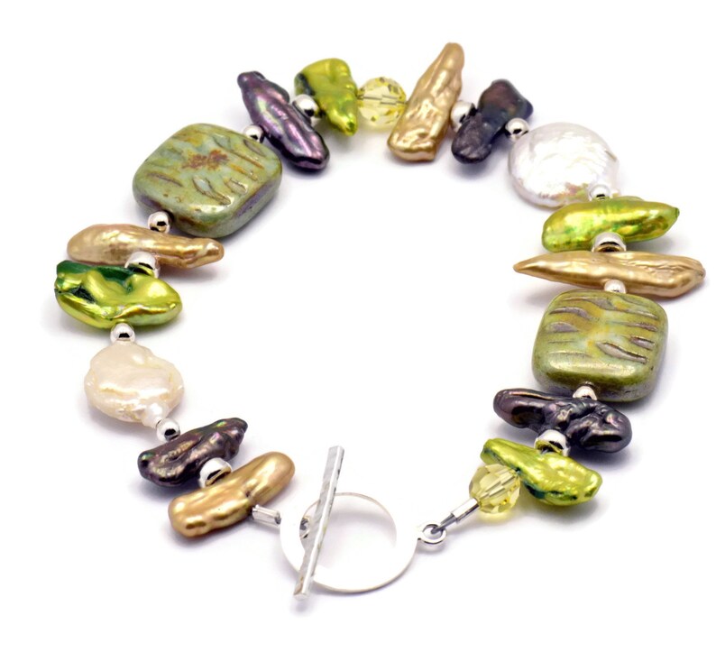 K&ouml;nnte beinhalten: Ein Armband aus gr&uuml;nen, goldenen und violetten S&uuml;&szlig;wasserperlen mit einem silbernen Schiebeverschluss. Das Armband verf&uuml;gt &uuml;ber eine Vielzahl von Formen und Gr&ouml;&szlig;en von Perlen, darunter runde, ovale und rechteckige.
