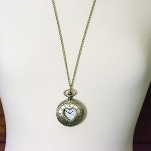 Pode incluir: Um colar dourado com um pingente de rel&oacute;gio de bolso em forma de cora&ccedil;&atilde;o. O mostrador do rel&oacute;gio tem algarismos romanos e uma janela em forma de cora&ccedil;&atilde;o.