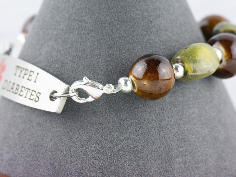 May include: A silver bracelet with a tag that reads "Type 1 Diabetes". The bracelet is made with brown and green ceramic beads and a silver clasp.