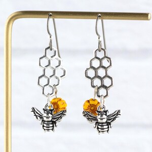 May include: Silver-tone bee earrings with honeycomb-shaped details. Each earring has a detailed bee charm, a yellow bead, and a honeycomb design. The earrings are displayed on a gold-coloured stand.
