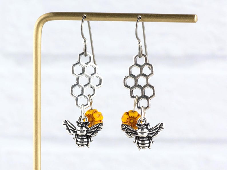 May include: Silver-tone bee earrings with honeycomb-shaped details. Each earring has a detailed bee charm, a yellow bead, and a honeycomb design. The earrings are displayed on a gold-coloured stand.