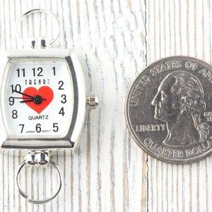 Puede incluir: Un reloj de cuarzo plateado con esfera blanca, n&uacute;meros negros y un coraz&oacute;n rojo. El reloj tiene anillos para sujetarlo. Una moneda de 25 centavos estadounidense est&aacute; al lado del reloj para comparar el tama&ntilde;o. El reloj mide unos 3,8 cm.