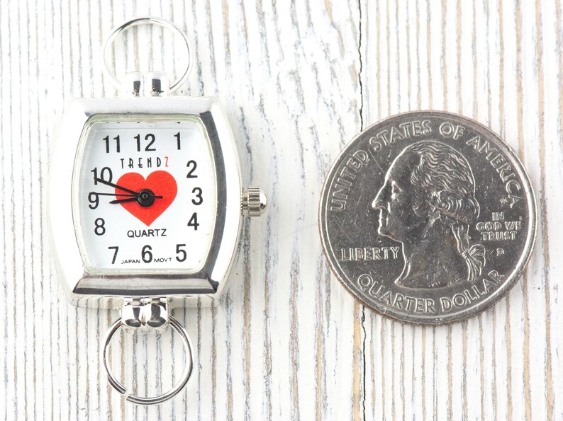 Puede incluir: Un reloj de cuarzo plateado con esfera blanca, n&uacute;meros negros y un coraz&oacute;n rojo. El reloj tiene anillos para sujetarlo. Una moneda de 25 centavos estadounidense est&aacute; al lado del reloj para comparar el tama&ntilde;o. El reloj mide unos 3,8 cm.