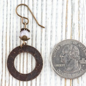 Puede incluir: Un solo pendiente de color bronce con un dise&ntilde;o circular y una peque&ntilde;a cuenta de color p&uacute;rpura claro. El pendiente est&aacute; junto a una moneda de veinticinco centavos estadounidense para comparar el tama&ntilde;o.