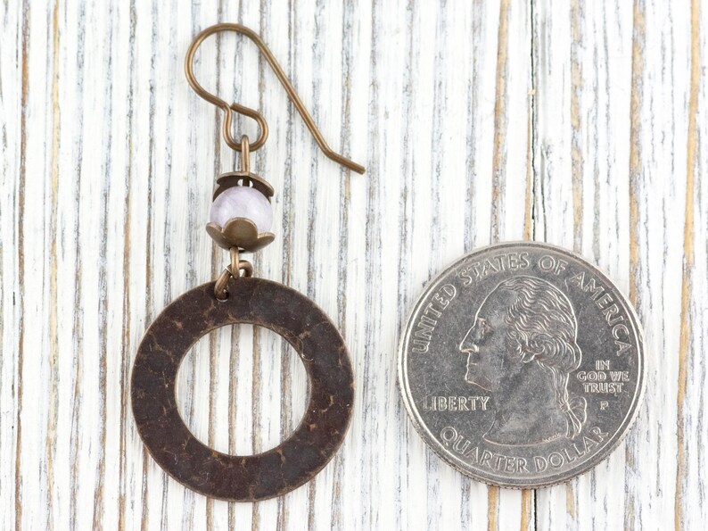 Puede incluir: Un solo pendiente de color bronce con un dise&ntilde;o circular y una peque&ntilde;a cuenta de color p&uacute;rpura claro. El pendiente est&aacute; junto a una moneda de veinticinco centavos estadounidense para comparar el tama&ntilde;o.
