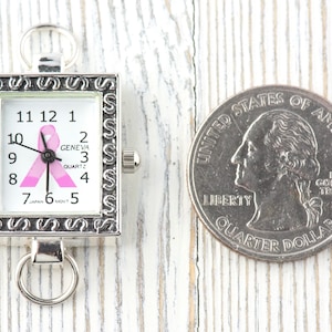 Op de afbeelding: Een kleine, vierkante horloge met een zilverkleurig frame en een witte wijzerplaat met een roze lintontwerp. De wijzerplaat toont de cijfers 1 tot en met 12. Een kwart dollar munt ligt naast het horloge.