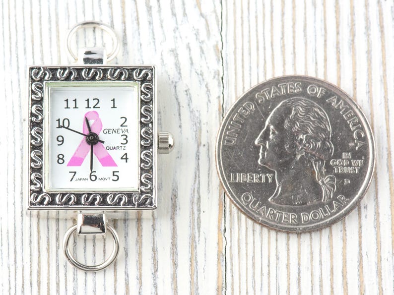 Op de afbeelding: Een kleine, vierkante horloge met een zilverkleurig frame en een witte wijzerplaat met een roze lintontwerp. De wijzerplaat toont de cijfers 1 tot en met 12. Een kwart dollar munt ligt naast het horloge.