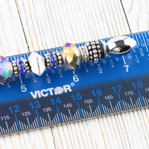 K&ouml;nnte beinhalten: Nahaufnahme eines silbernen Charm-Armbands mit verschiedenen Perlen, darunter eine klare, schillernde Perle, eine blaue Kristallperle, eine silberne Perle mit strukturierter Oberfl&auml;che, eine klare Kristallperle mit goldfarbenem Akzent, eine silberne Perle mit strukturierter Oberfl&auml;che und eine silberne Perle mit strukturierter Oberfl&auml;che. Das Armband wird auf einem blauen Lineal mit schwarzen Markierungen gemessen.